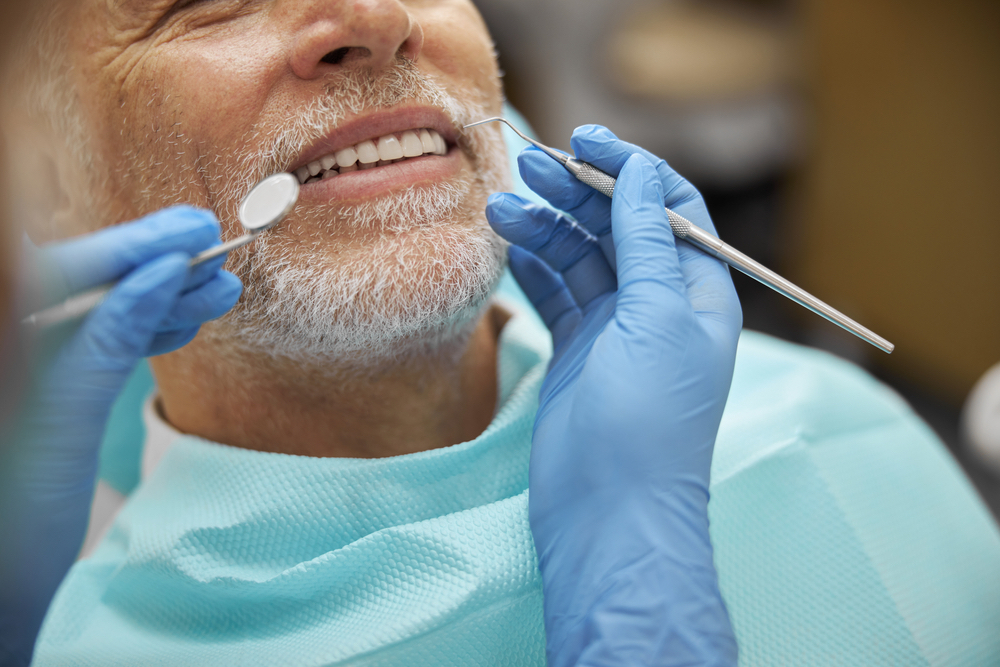 A cropped photo of a male senior smiling while having his teeth examined by a dentist.