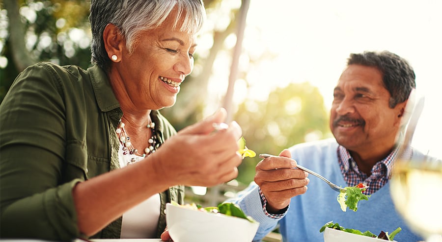 An older couple are both sitting at a table outside together smiling while eating salad.