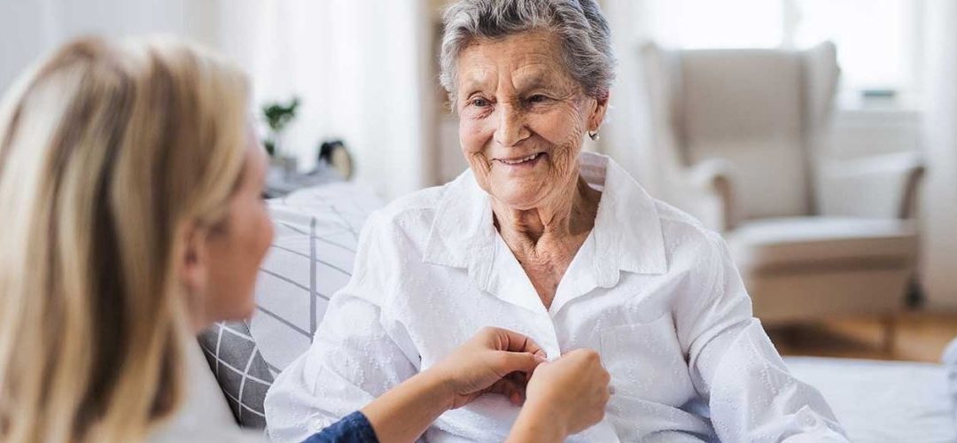 A caregiver is buttoning the top buttons of the shirt of an elderly woman as they are both smiling while talking.