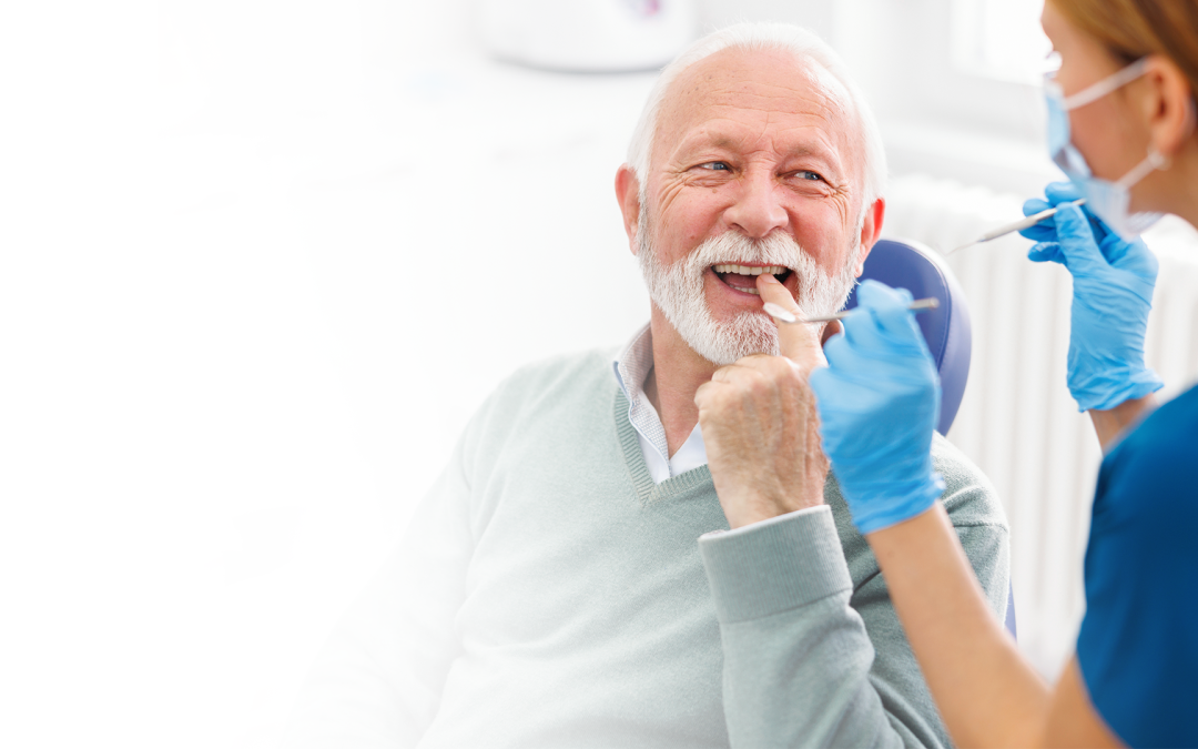 An elderly male is sitting in a chair smiling and pointing at his teeth while speaking with a Toothpicks staff member sitting beside him.