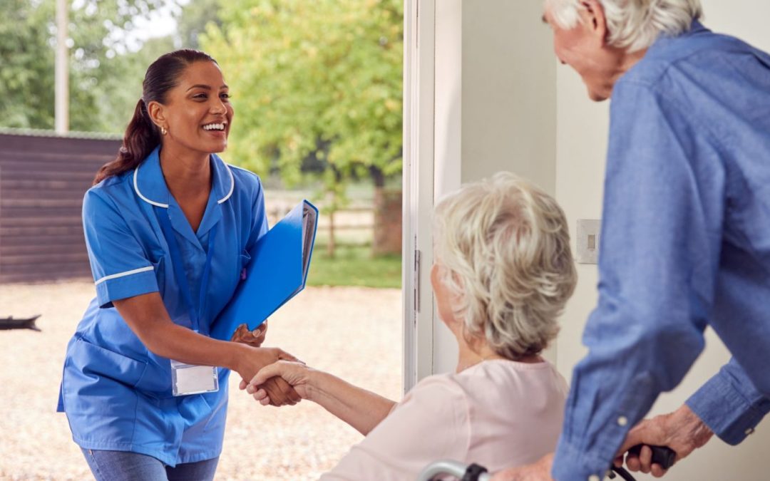 An elderly male is pushing a wheelchair with an elderly woman sitting in it, and they are greeting a Toothpicks staff member at their front door together. The Toothpicks staff member is shaking hands with the elderly woman in the wheelchair, and all 3 people are smiling.