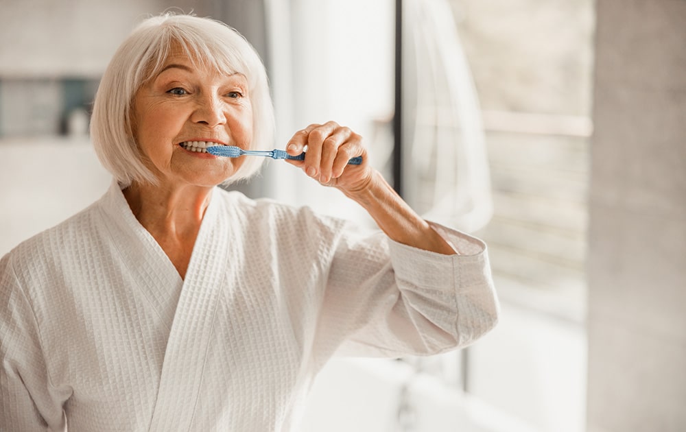 An elderly woman brushing her teeth in the bathroom mirror.